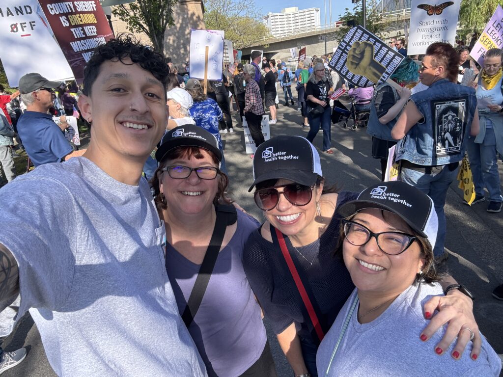 Four BHT staff members—Noah, Kim, Allison, and Angela—smile and pose for a selfie at a local outdoor rally. They are wearing Better Health Together (BHT) gear, including branded hats and shirts. Behind them, a diverse crowd holds up protest signs and banners, with trees and buildings visible in the background. The image has rounded corners with a thin purple border.