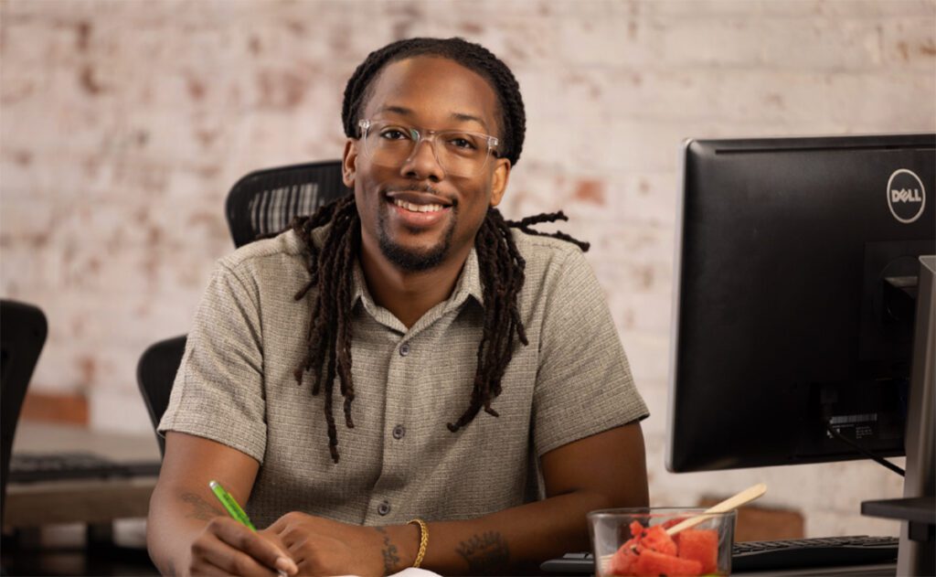A smiling person with long dreadlocks, glasses, and a short-sleeve button-up shirt sits at a desk in front of a computer monitor. They are holding a green pencil and appear to be working or studying. A cup with watermelon slices and chopsticks is visible on the desk. The background features a white brick wall.