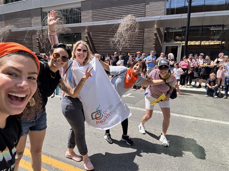5 BHT staff members of various heights, ages, and races walk down the street with a Better Health Together Sign to celebrate pride. The group is smiling