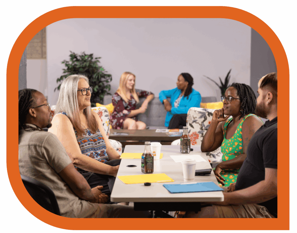 “A diverse group of adults sits around tables in conversation during a community meeting. In the background, two pairs of people talk on couches. The atmosphere is warm and collaborative, with notebooks, drinks, and folders on the table. The image is framed by a rounded orange border.”