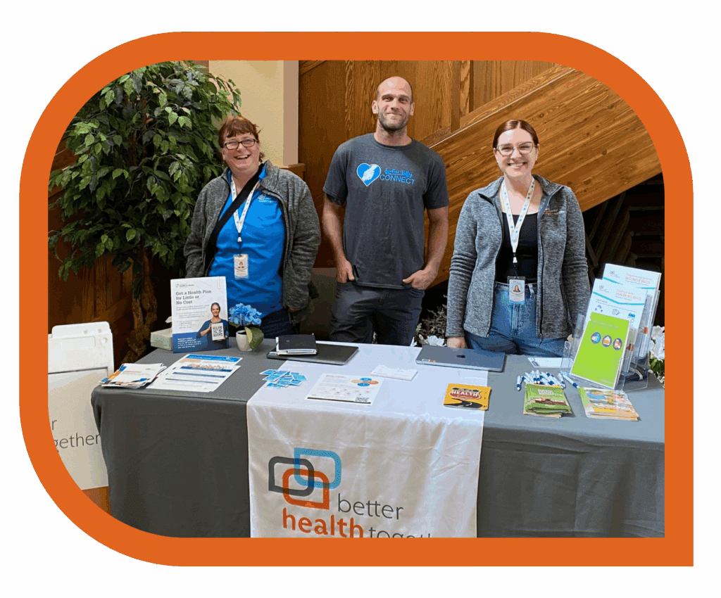 Three smiling BHT Staff—Kim, Ryan, and Sara—stand behind a resource table covered with brochures, pens, flyers, and health-related materials. The table displays a "Better Health Together" banner. Behind them is a wood-paneled wall and a potted plant. The photo is framed with rounded corners and an orange border.